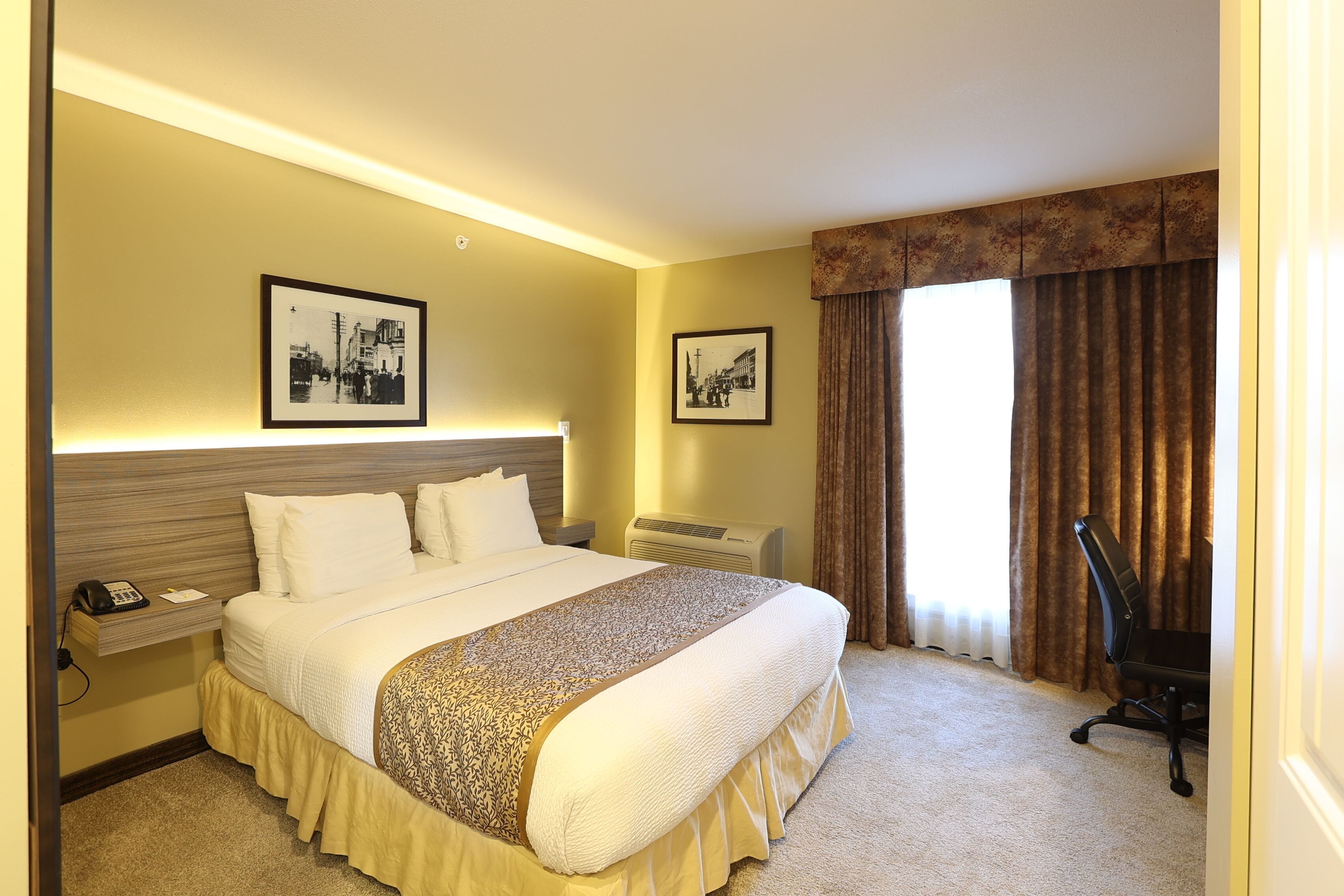 Habitación de hotel con una cama grande, una mesita de noche con teléfono, dos cuadros en la pared, una ventana con cortinas y una silla.