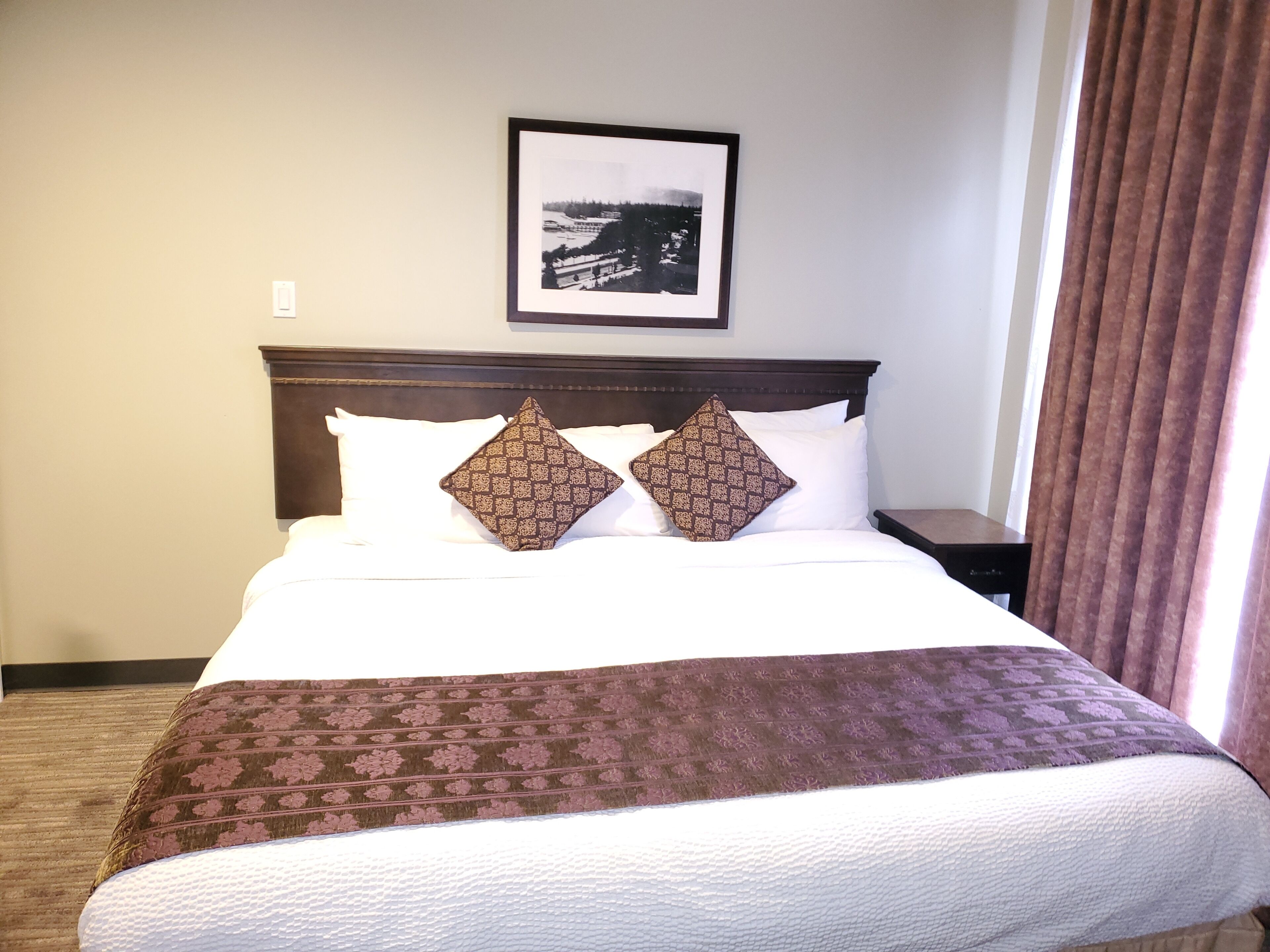 Una habitación de hotel con una cama bien hecha, un cuadro enmarcado en la pared y una ventana con cortinas.