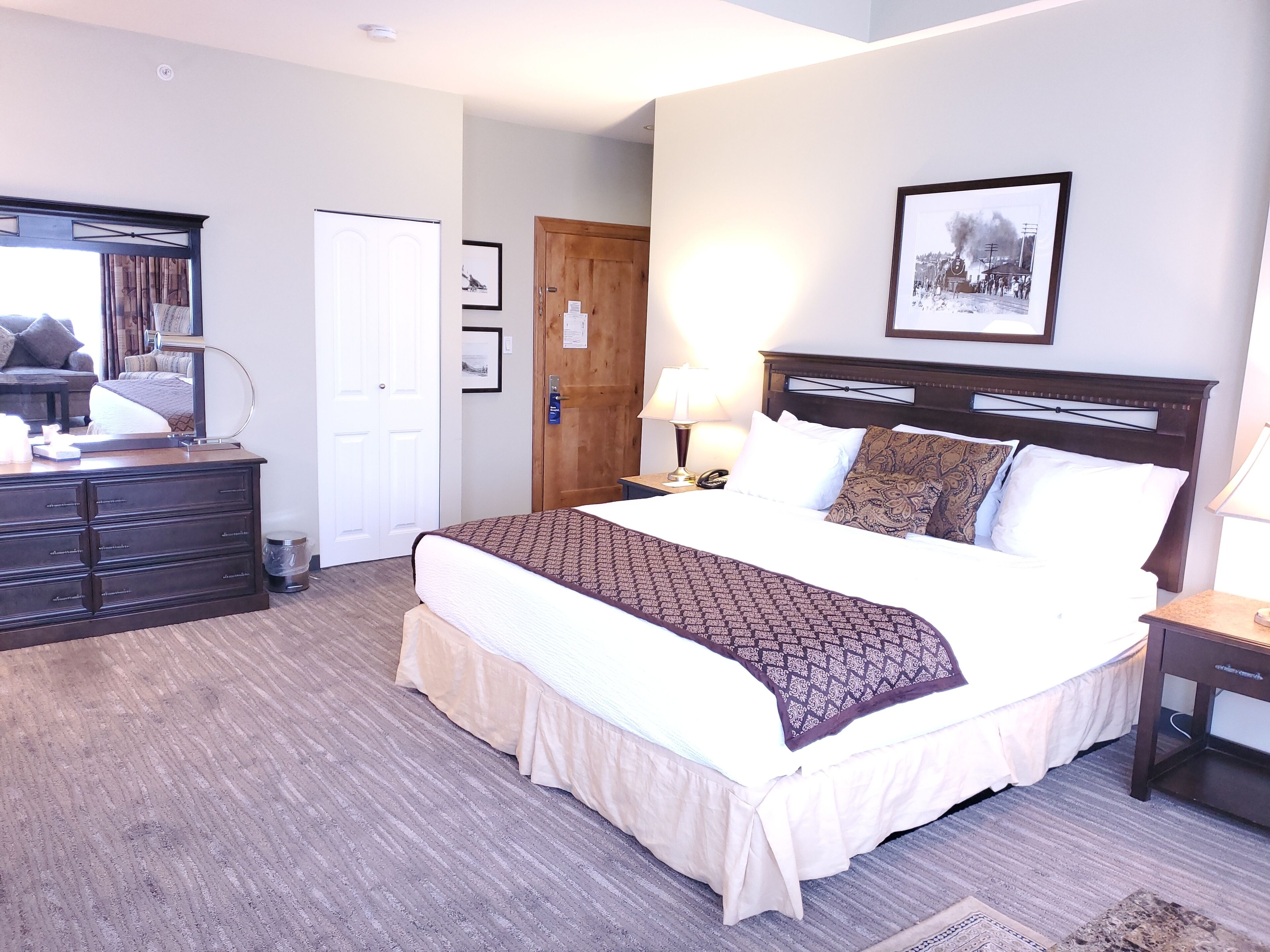 A neatly arranged bedroom with a large bed, a dresser, a bedside table with a lamp, and a framed picture on the wall.