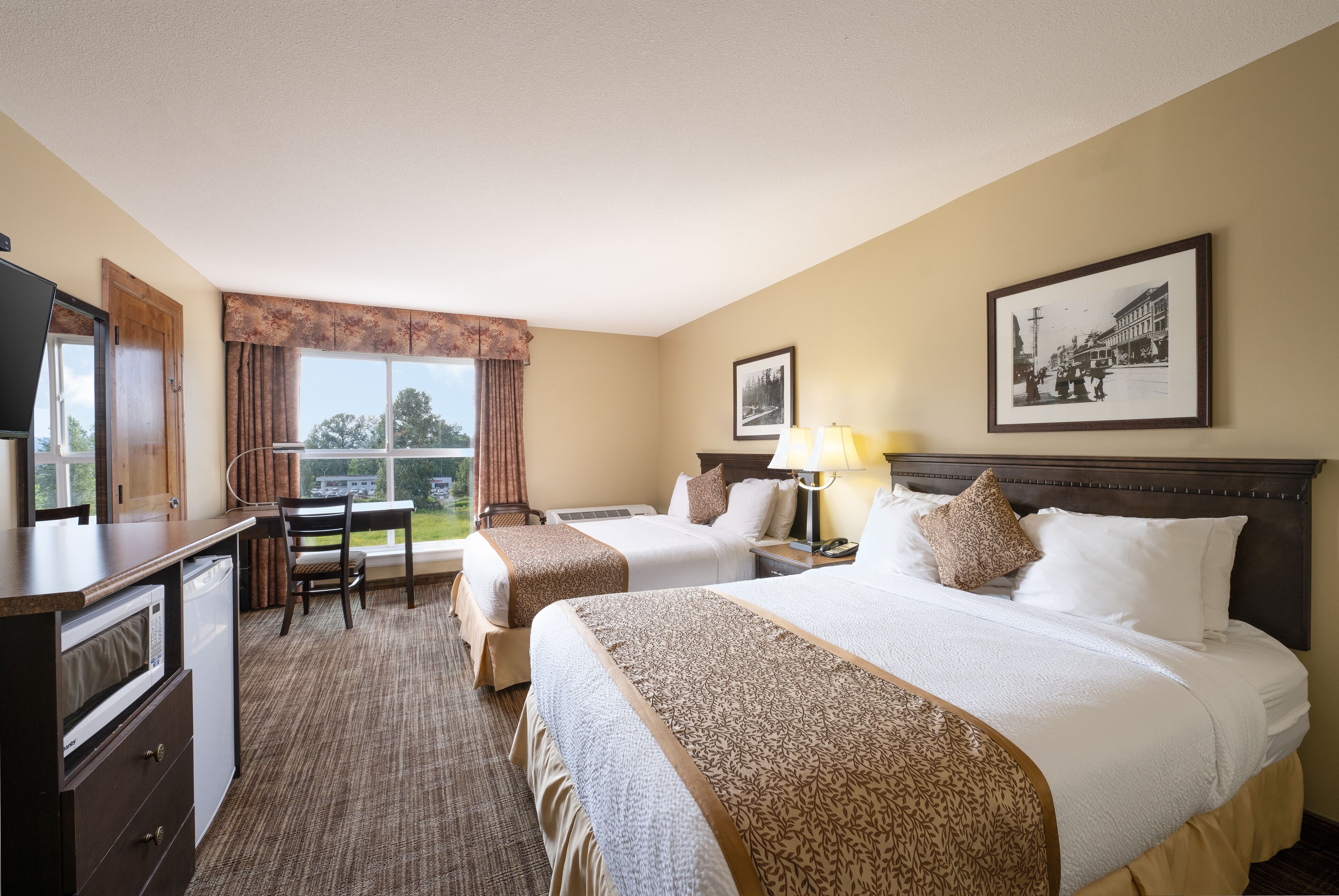 A hotel room with two beds, a desk, a microwave, and a window with a view of the outdoors.