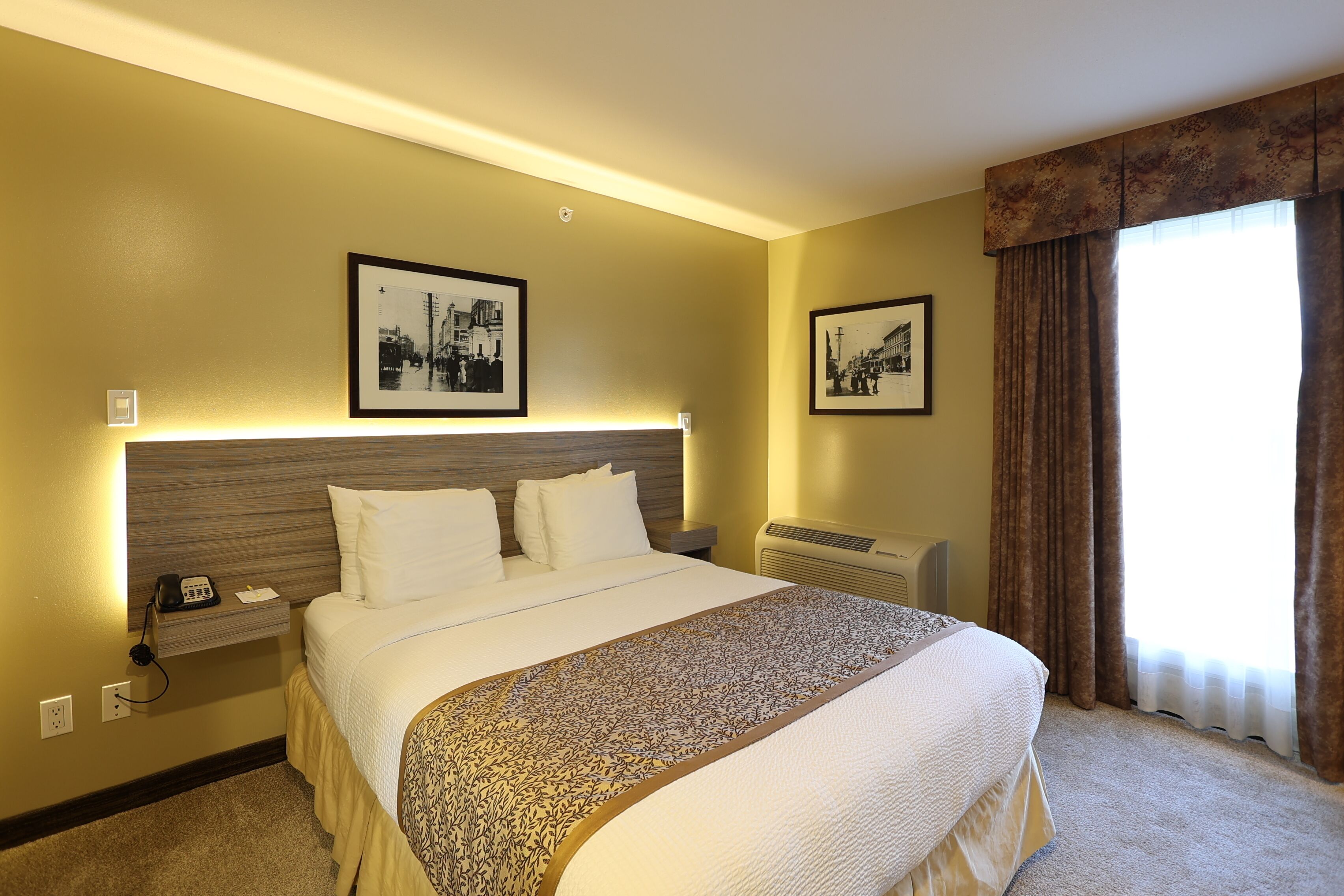A hotel room with a large bed, two framed pictures on the wall, a bedside table with a phone, and a window with curtains.