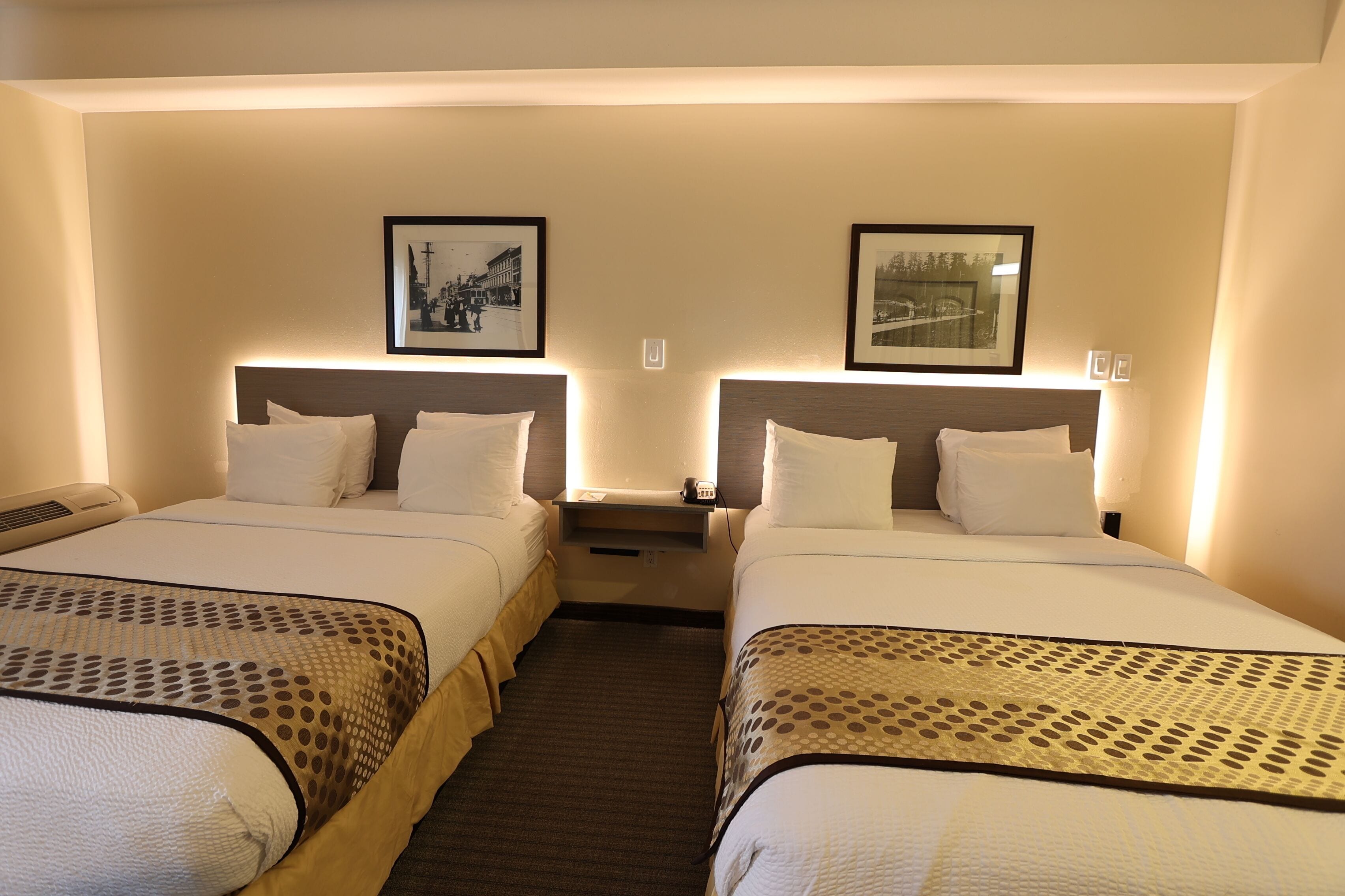 A hotel room with two beds, each with a headboard, a bedside table, and a wall-mounted phone. There are framed pictures above each bed.