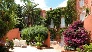 Un patio con suelo de ladrillo, mobiliario exterior blanco y abundante vegetación, incluyendo una palmera y plantas con flores.