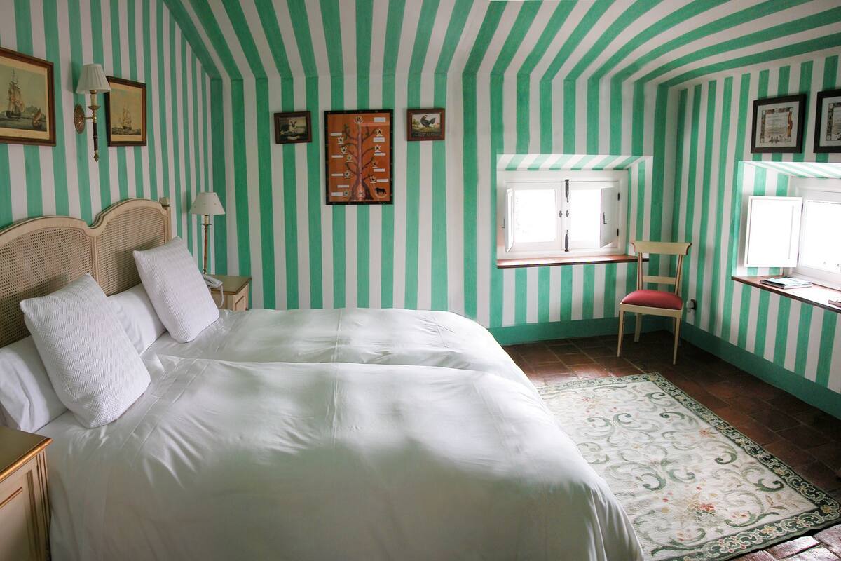 A room with striped green and white walls, a bed with white linens, a wooden headboard, a chair with a red cushion, and a window with a view of the outside.