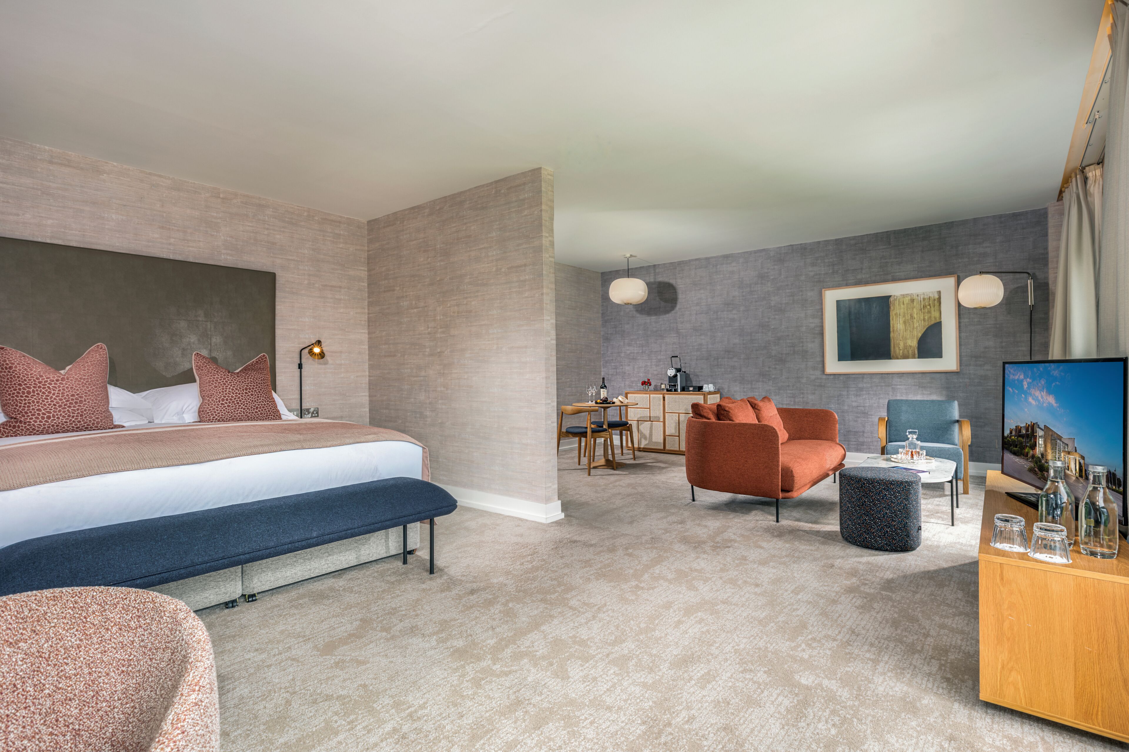 A modern hotel room with a large bed, a seating area with a sofa and armchair, a dining table, and a TV.