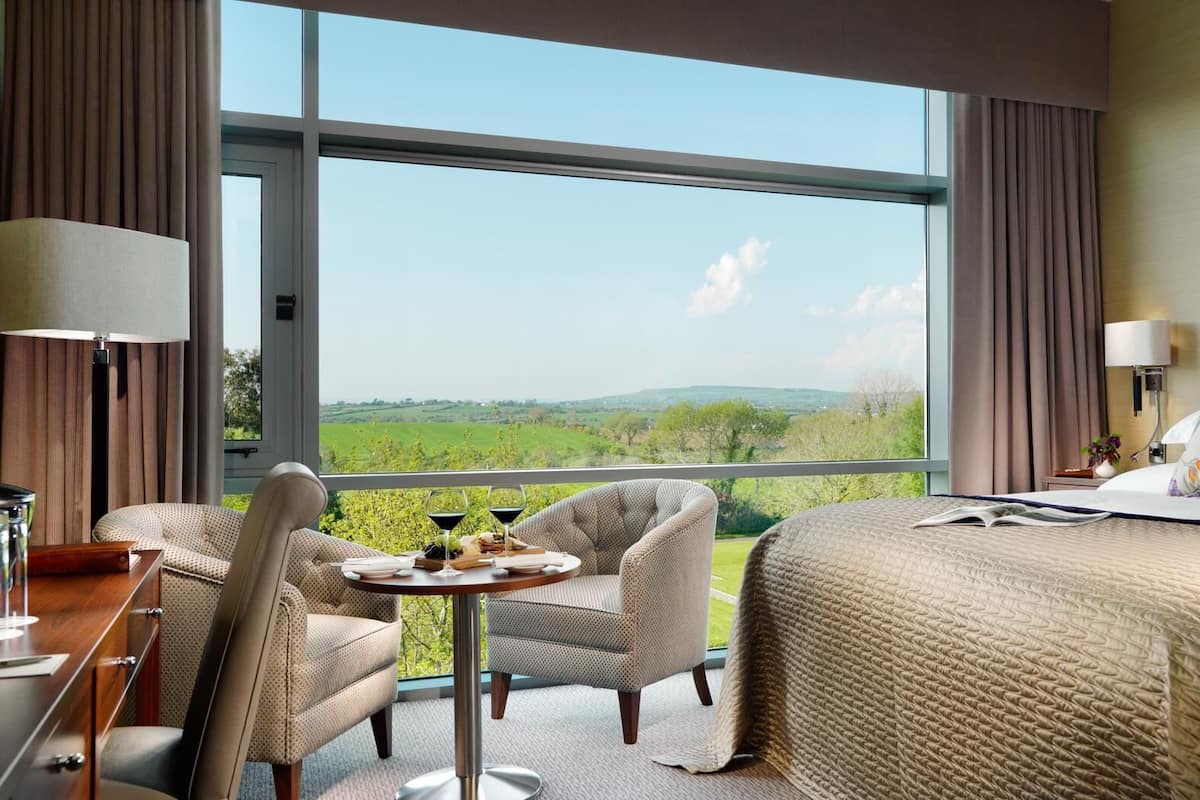Ein Hotelzimmer mit großem Fenster, einem Bett, einem Schreibtisch mit Lampe, zwei Stühlen und einem kleinen Tisch mit Tablett voller Speisen und Getränke.