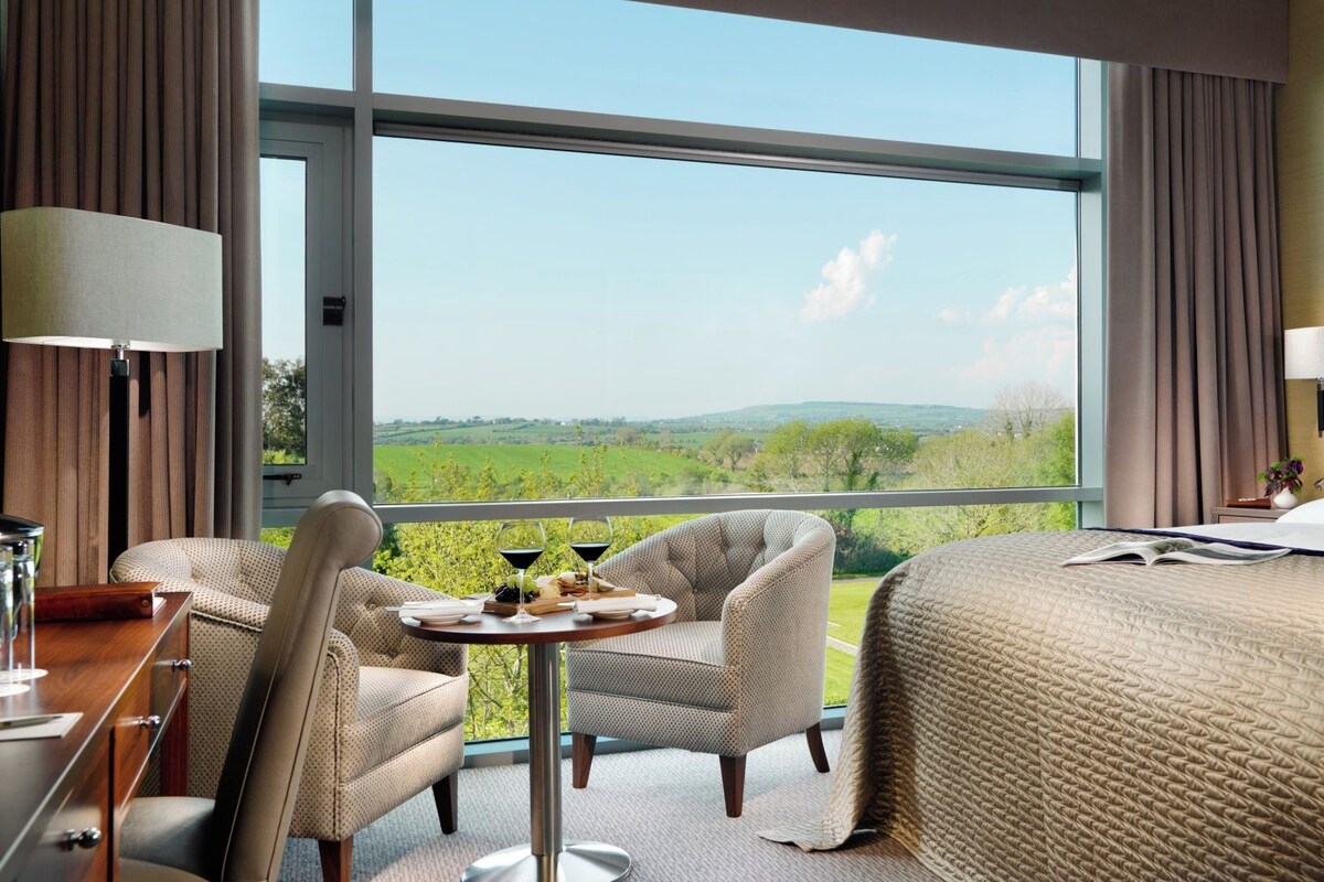 A hotel room with a large window, a bed, a dining area with two chairs, and a table with a bottle and glasses.