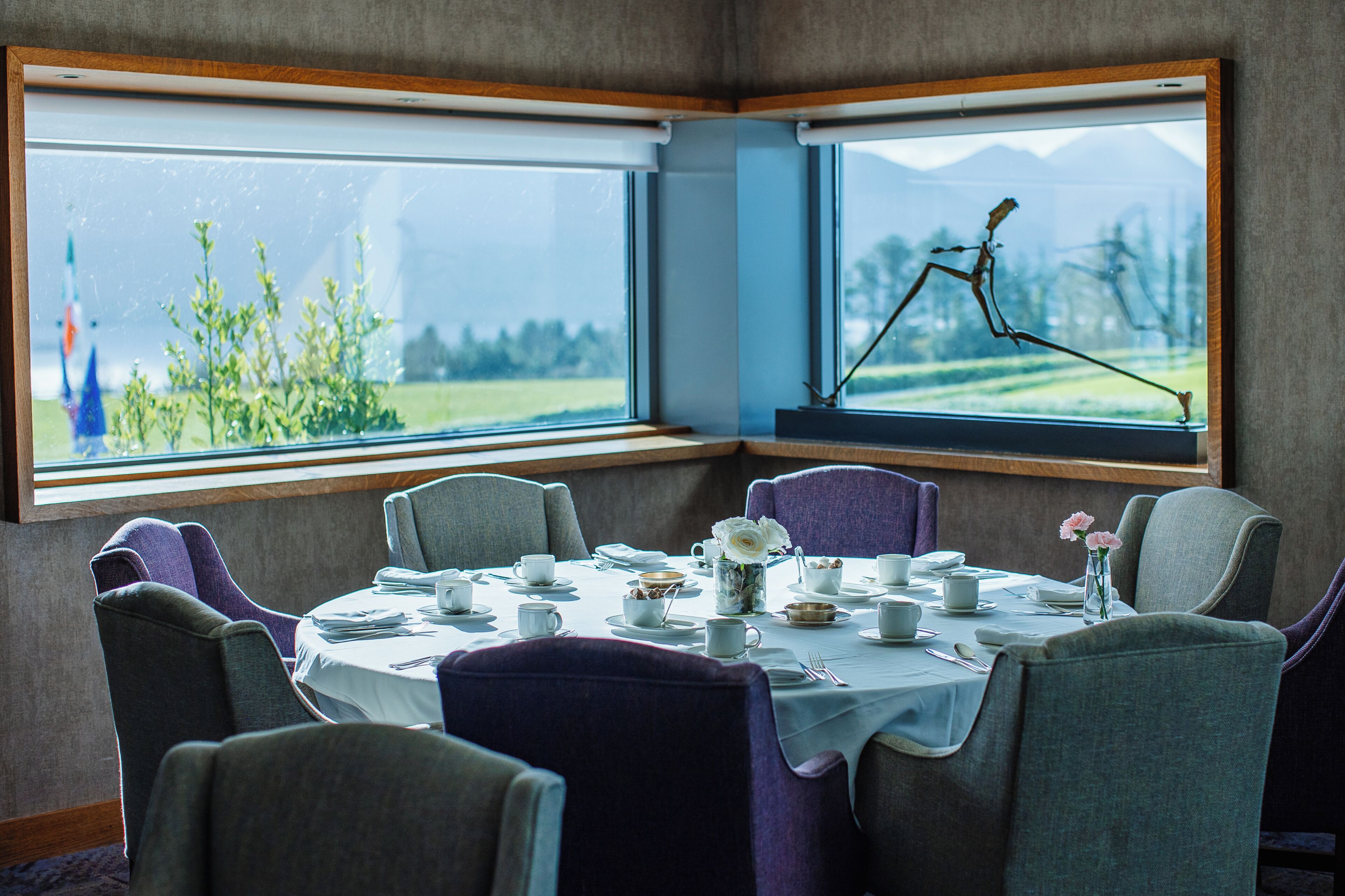 Ein Essbereich mit einem Tisch für vier Personen, Stühlen, einem Fenster mit Blick auf Grünflächen und Berge sowie einem dekorativen Insekt auf der Fensterbank.