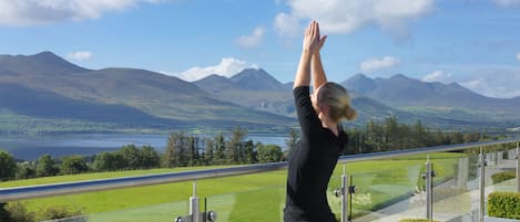 A person performing a yoga pose on a balcony with a scenic view of mountains and a lake.