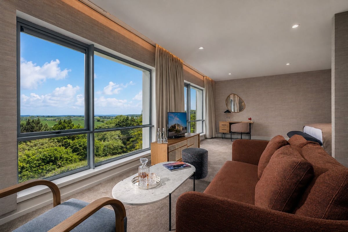 Ein modernes Hotelzimmer mit großen Fenstern, einer Couch, einem kleinen Tisch, einem Fernseher und einem Schreibtisch.