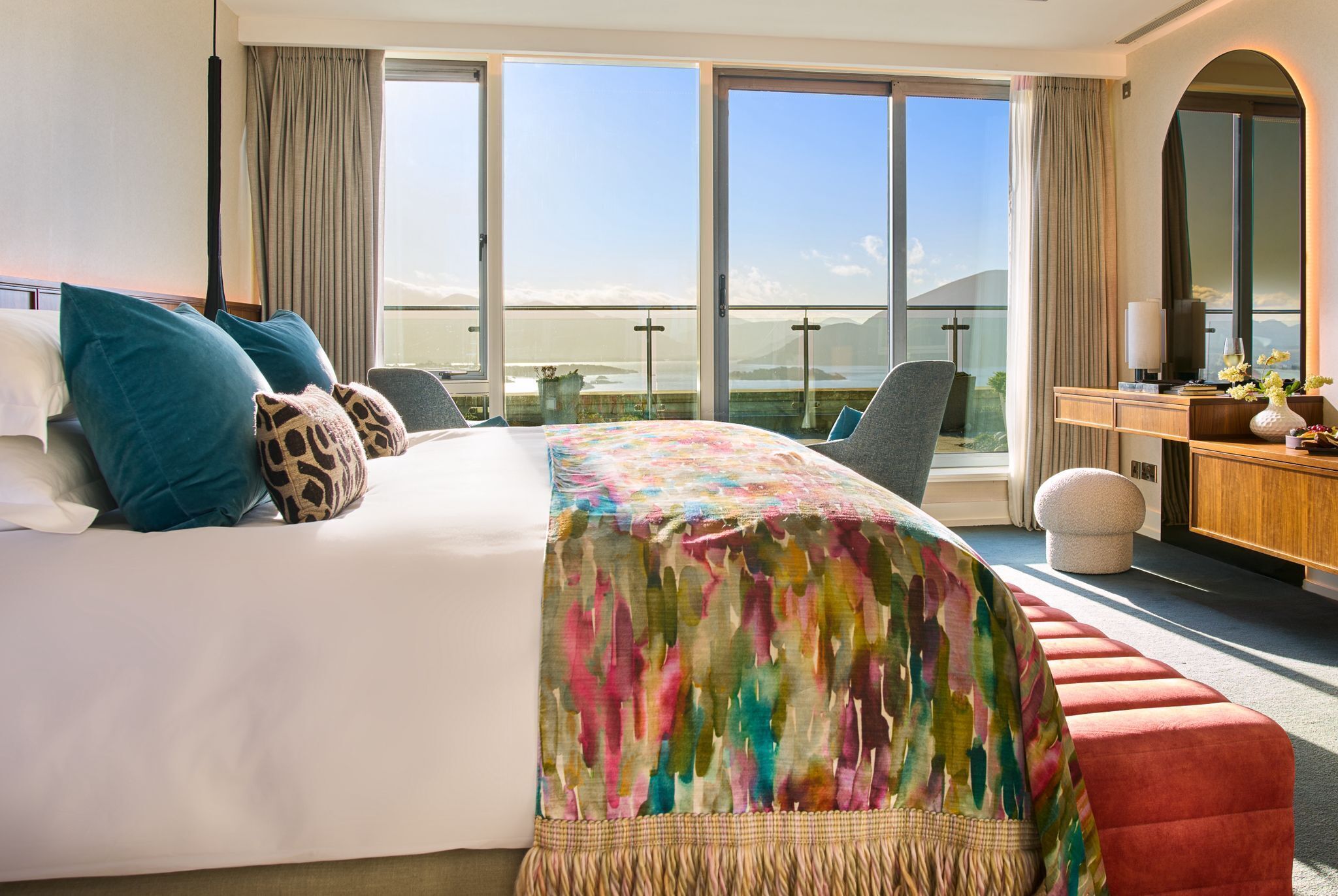 A hotel room with a large bed, a desk, a chair, and a view of the sea through large windows.