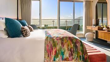 Une chambre d’hôtel avec un grand lit, un bureau, une chaise et une vue sur la mer grâce à de grandes fenêtres.