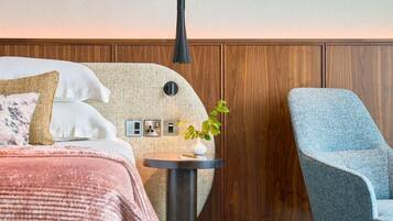 Quarto de hotel moderno com uma cama, uma poltrona cinza, uma pequena mesa com uma planta e lambris de madeira na parede.