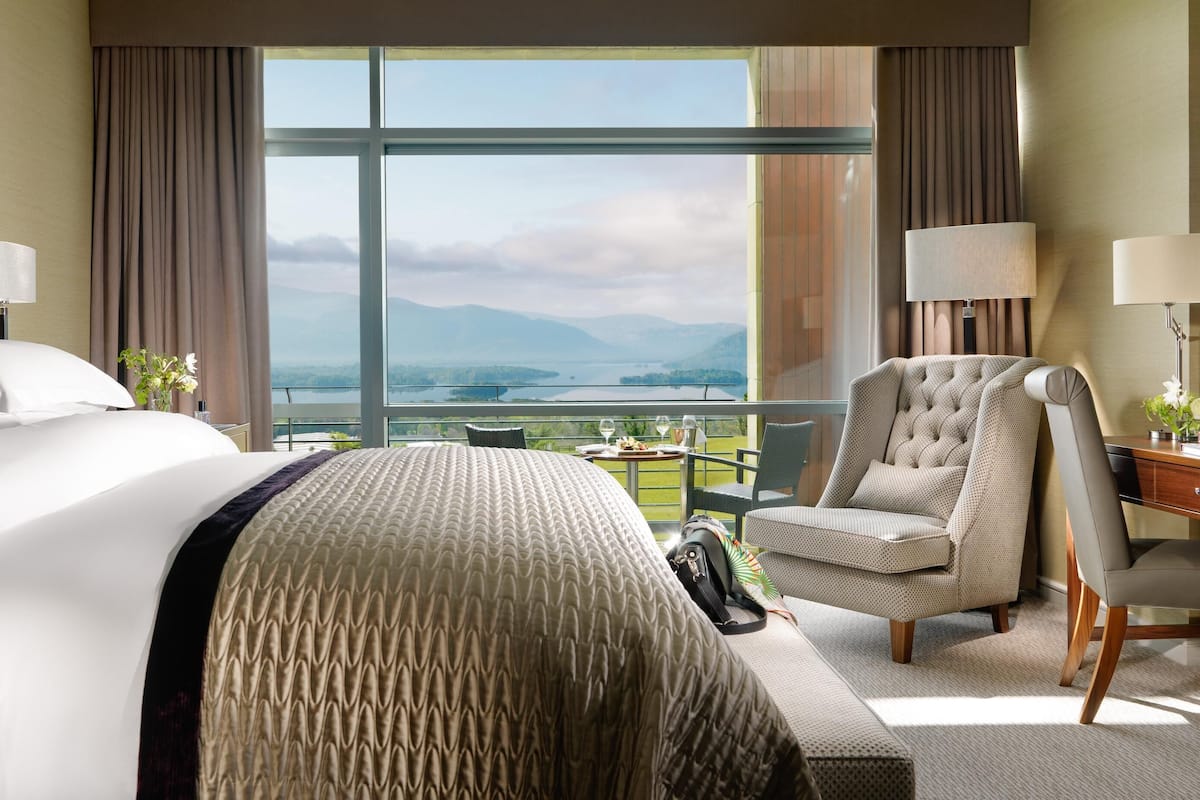 Ein Hotelzimmer mit einem großen Bett, einem Sessel, einem Schreibtisch und Blick auf einen See und Berge.