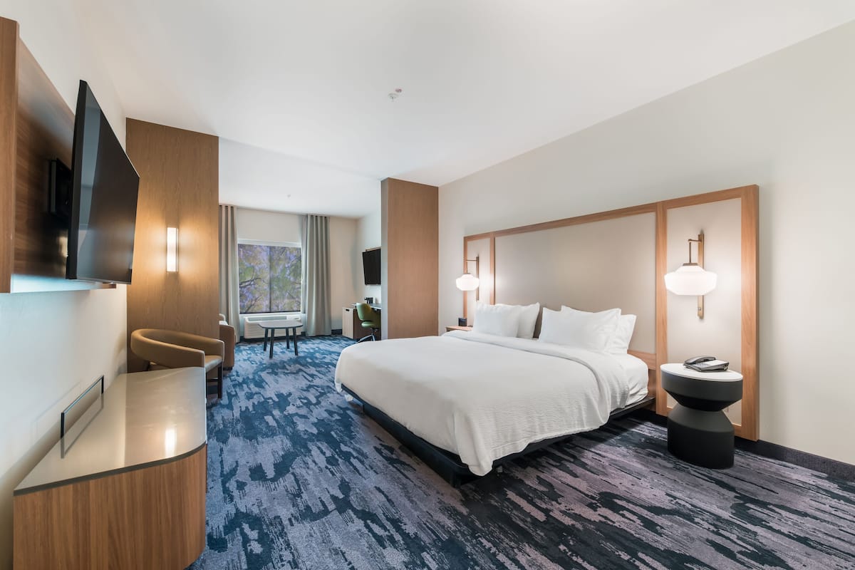 A hotel room with a large bed, a desk with a flat-screen TV, a seating area with a chair, and a large window.