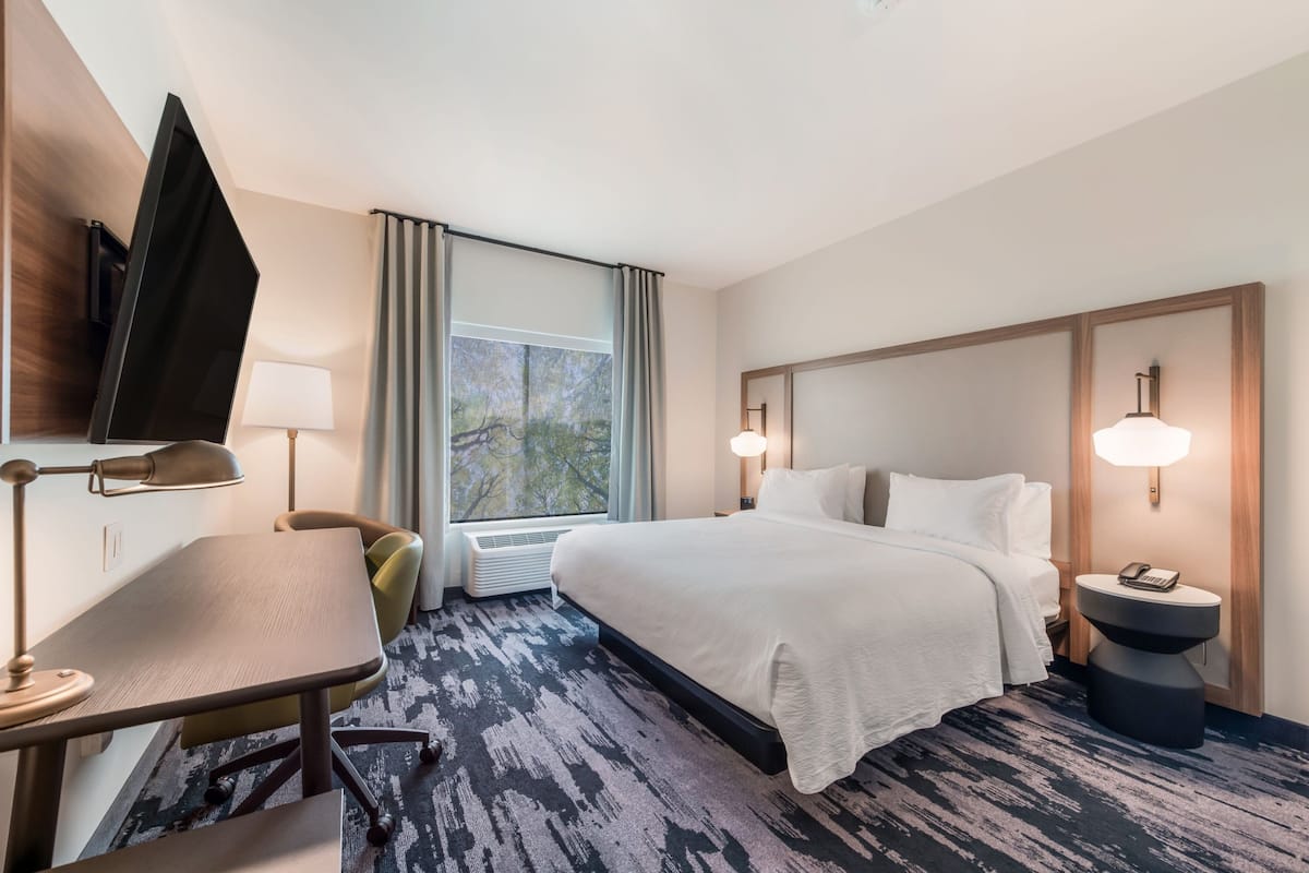 A hotel room with a bed, a desk, a chair, a TV, a lamp, and a window with curtains.