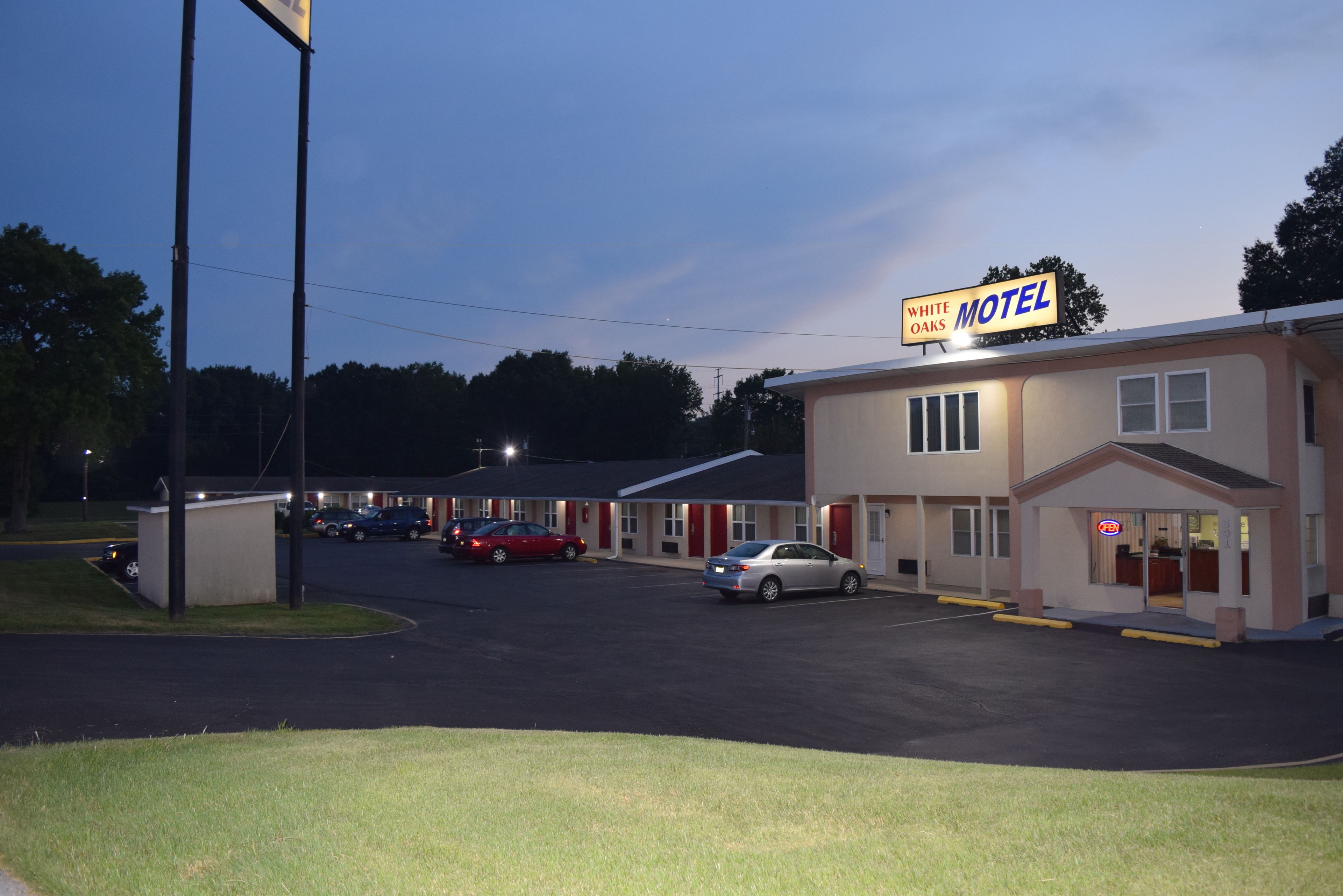 A motel with a sign reading "White Oaks Motel," featuring a parking lot with several cars, a small building to the left, and a visible entrance with a glass door.