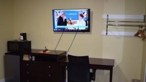 Una habitación con escritorio, silla, televisor montado en la pared, refrigerador pequeño y perchero.
