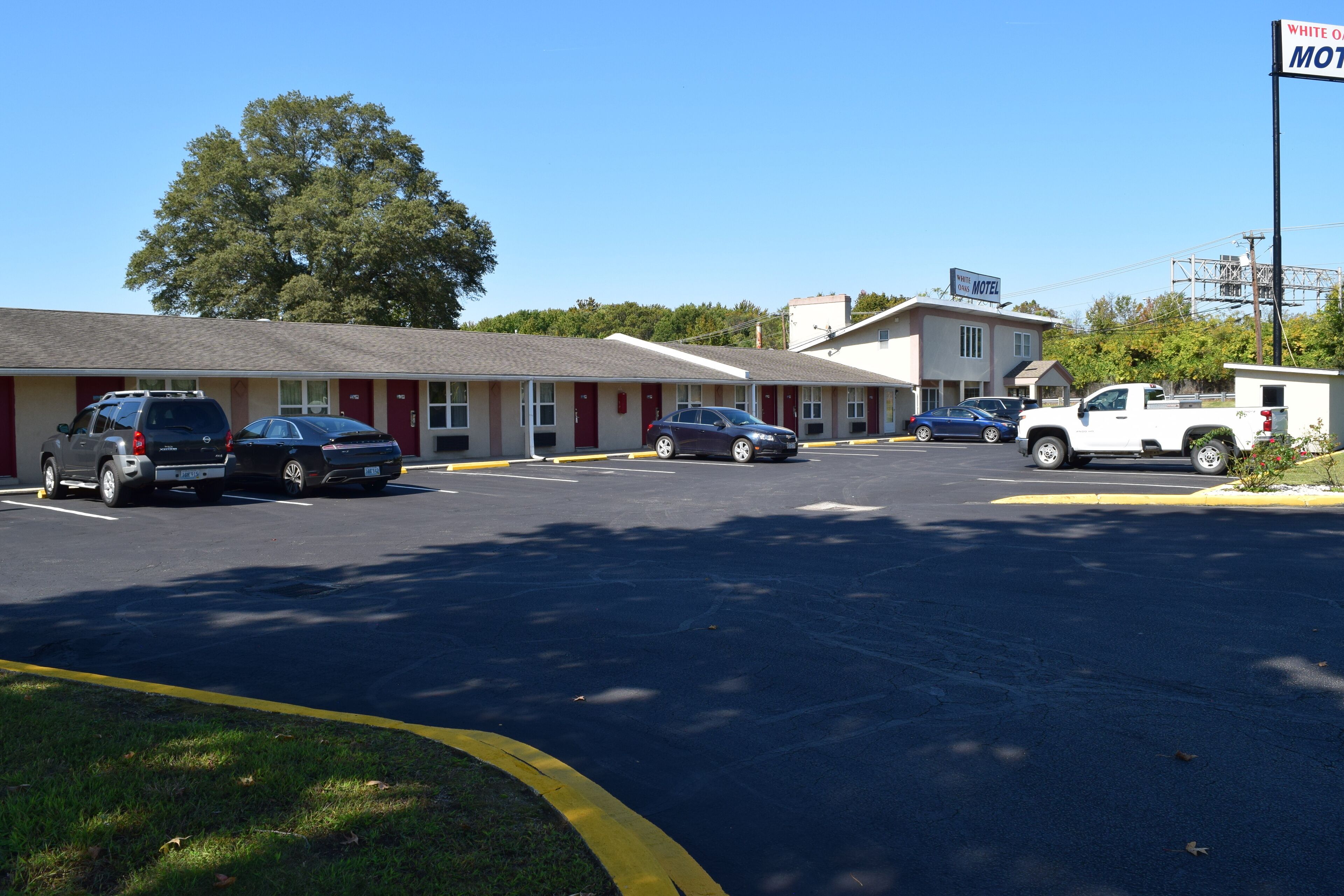 Un motel con una hilera de habitaciones, un estacionamiento y un árbol grande al fondo.