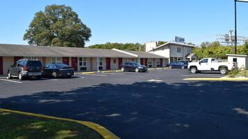 Un motel con una hilera de habitaciones, un estacionamiento y un árbol grande al fondo.