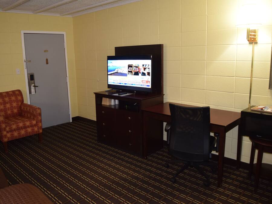 A hotel room with a television on a wooden stand, a patterned armchair, a desk with a chair, and a door with a keypad lock.