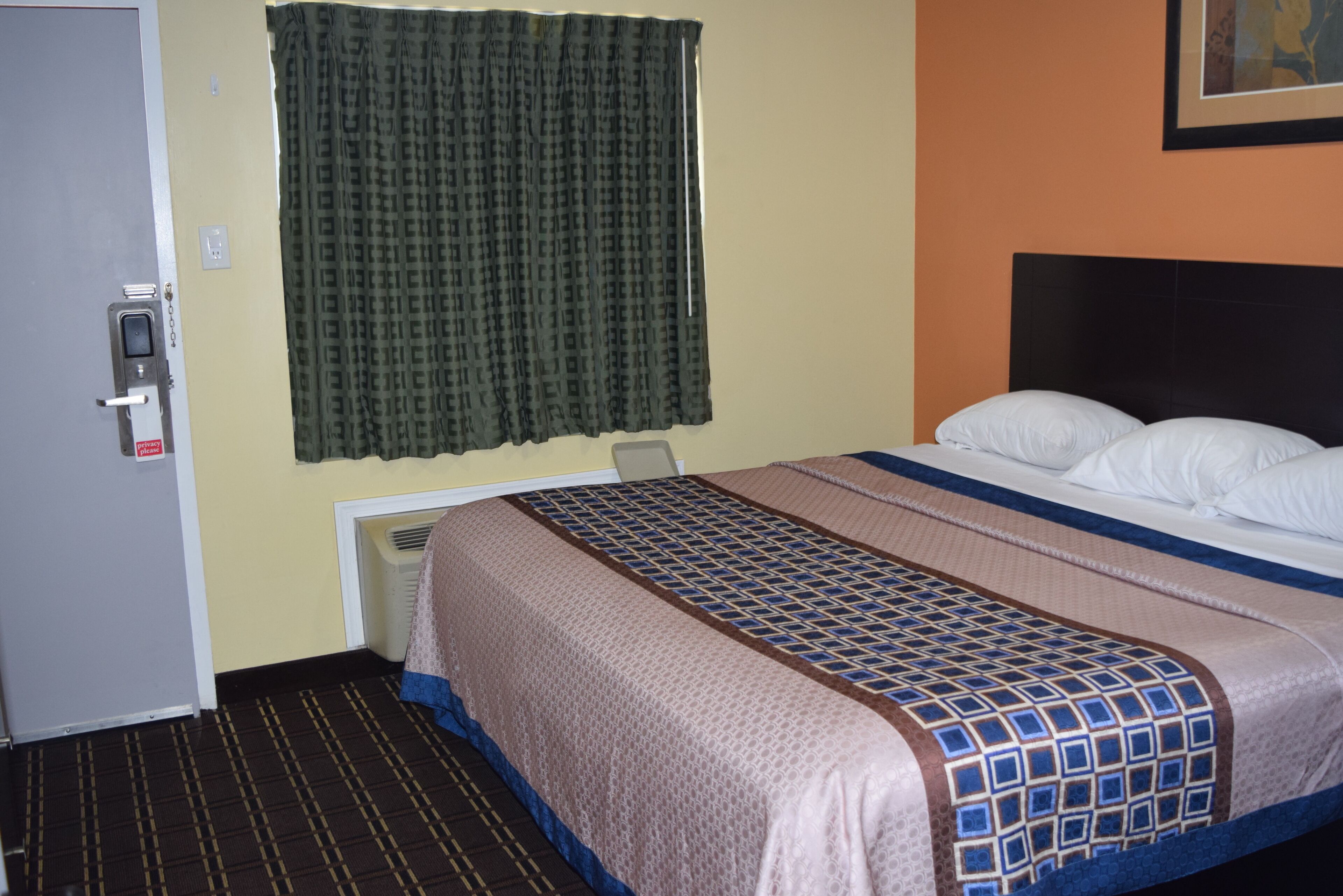 Habitación de hotel con cama, puerta, ventana con cortinas y un conducto de ventilación en la pared.