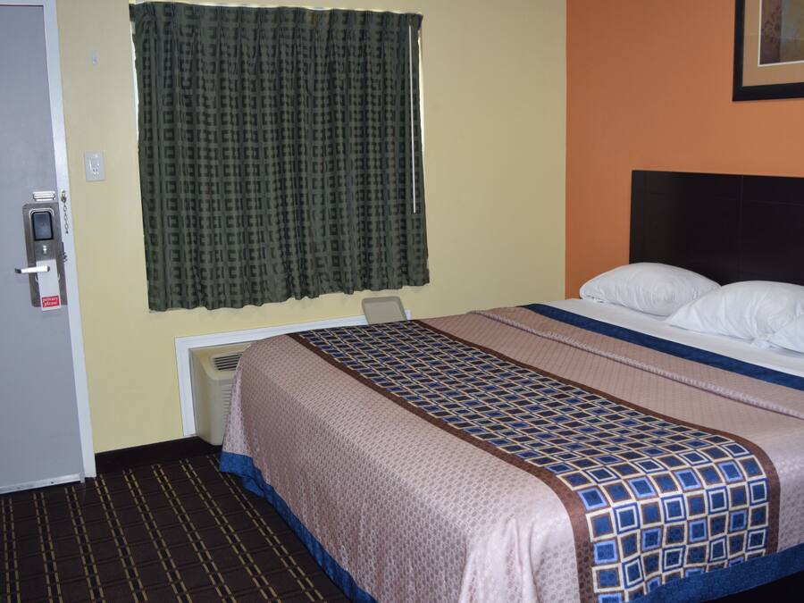 Habitación de hotel con cama, puerta, ventana con cortinas y un conducto de ventilación en la pared.