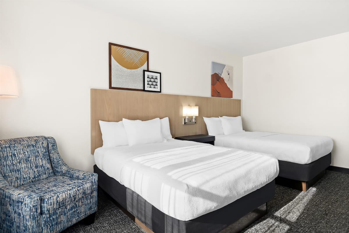 Quarto de hotel com duas camas, uma poltrona azul e obras de arte nas paredes.