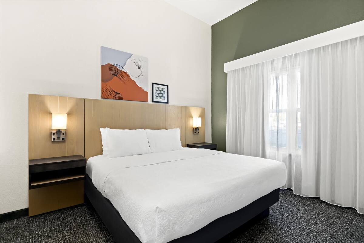 Quarto de hotel com uma cama grande, duas mesas de cabeceira, uma obra de arte na parede e uma janela com cortinas translúcidas.