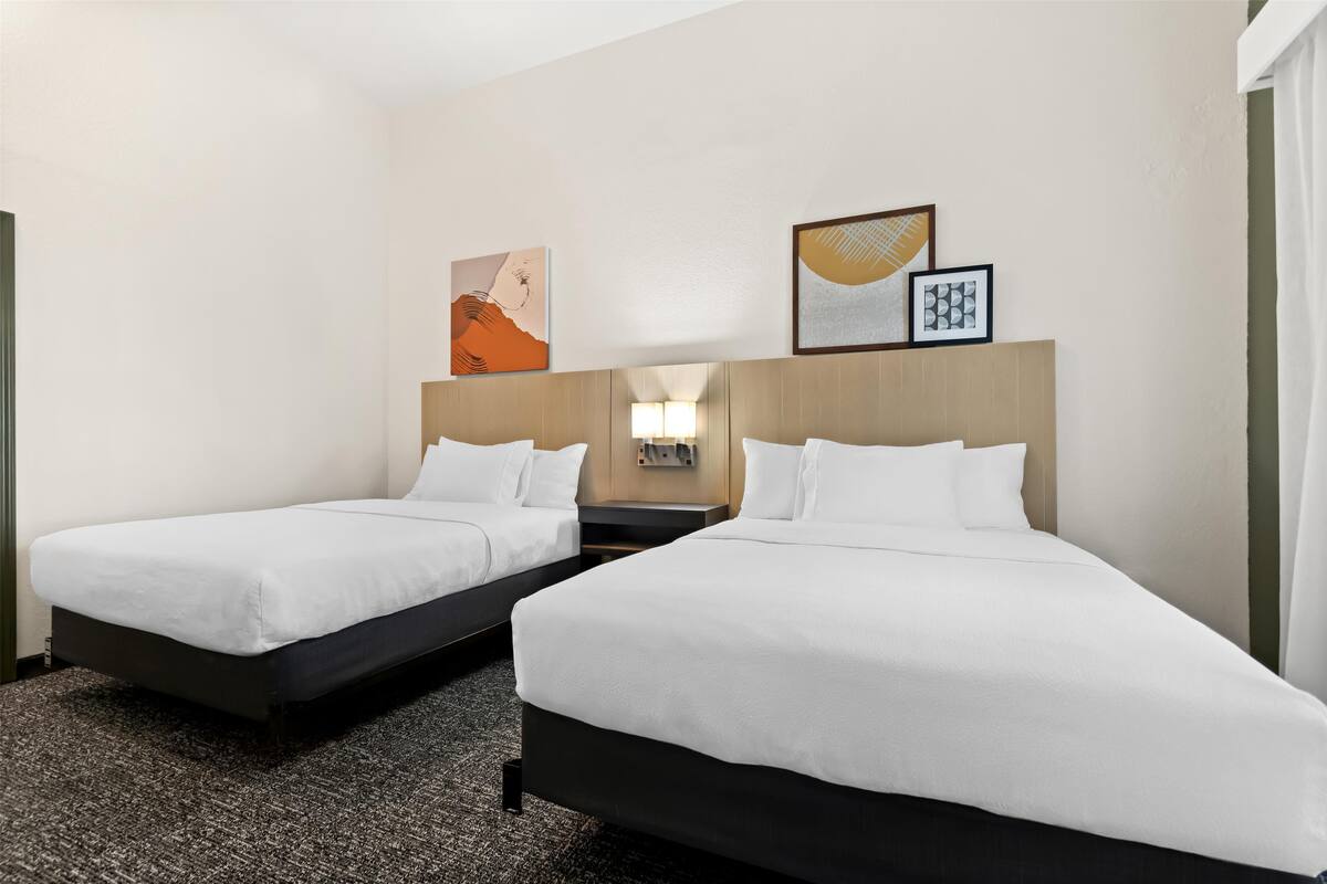 Quarto de hotel com duas camas de solteiro, uma mesa de cabeceira, um abajur e uma obra de arte na parede.