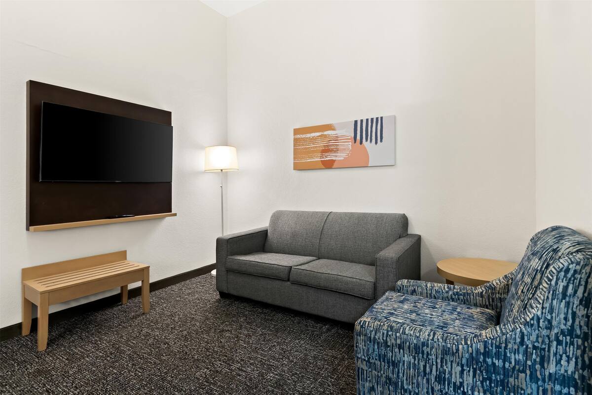 Um quarto com um sofá cinzento, uma poltrona azul estampada, um banco de madeira, uma televisão de tela plana na parede e um abajur.