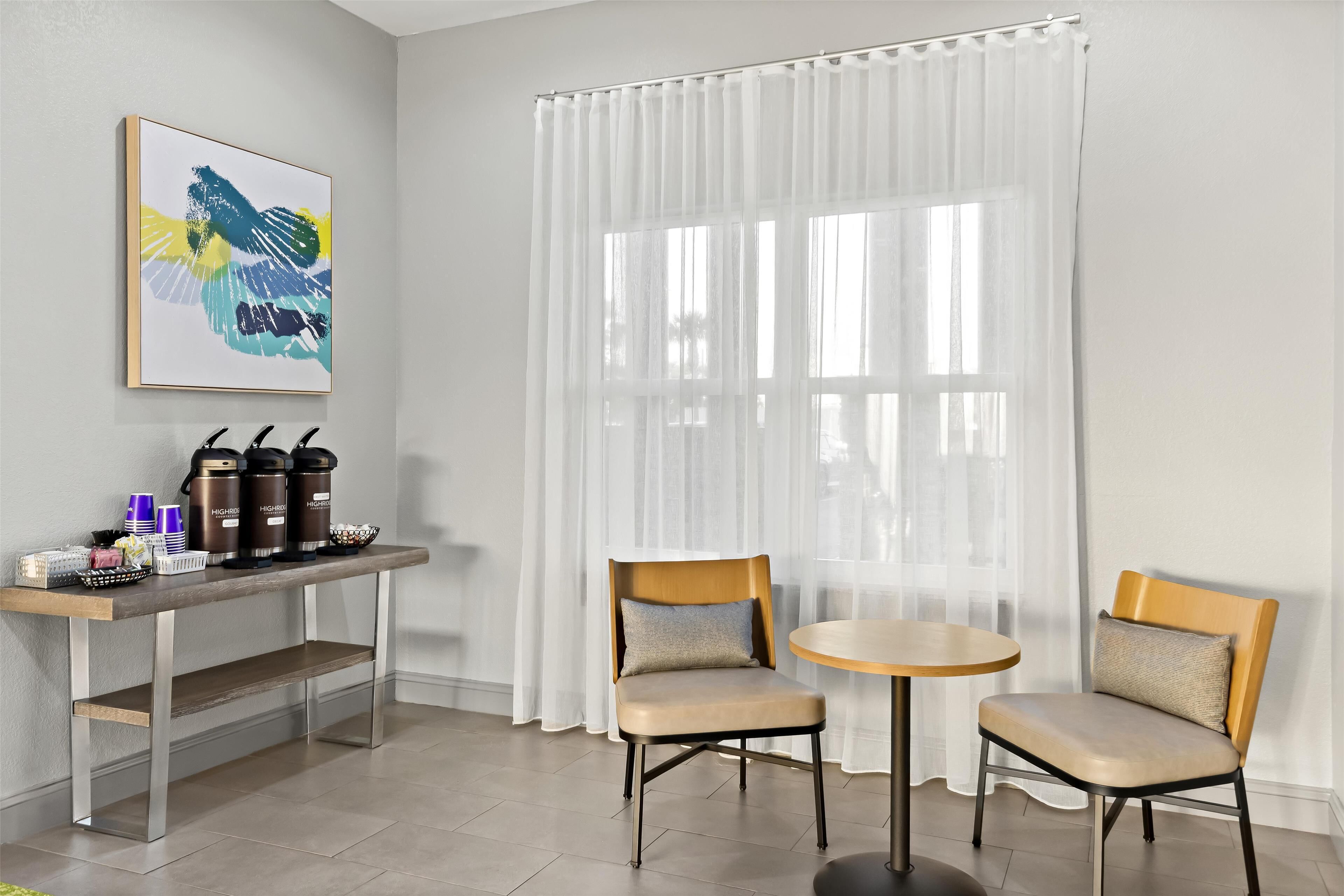 Una habitación moderna con dos sillas, una mesita, un espacio para café y un ventanal con cortinas translúcidas.