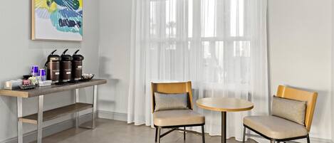 Una habitación moderna con dos sillas, una mesita, un espacio para café y un ventanal con cortinas translúcidas.