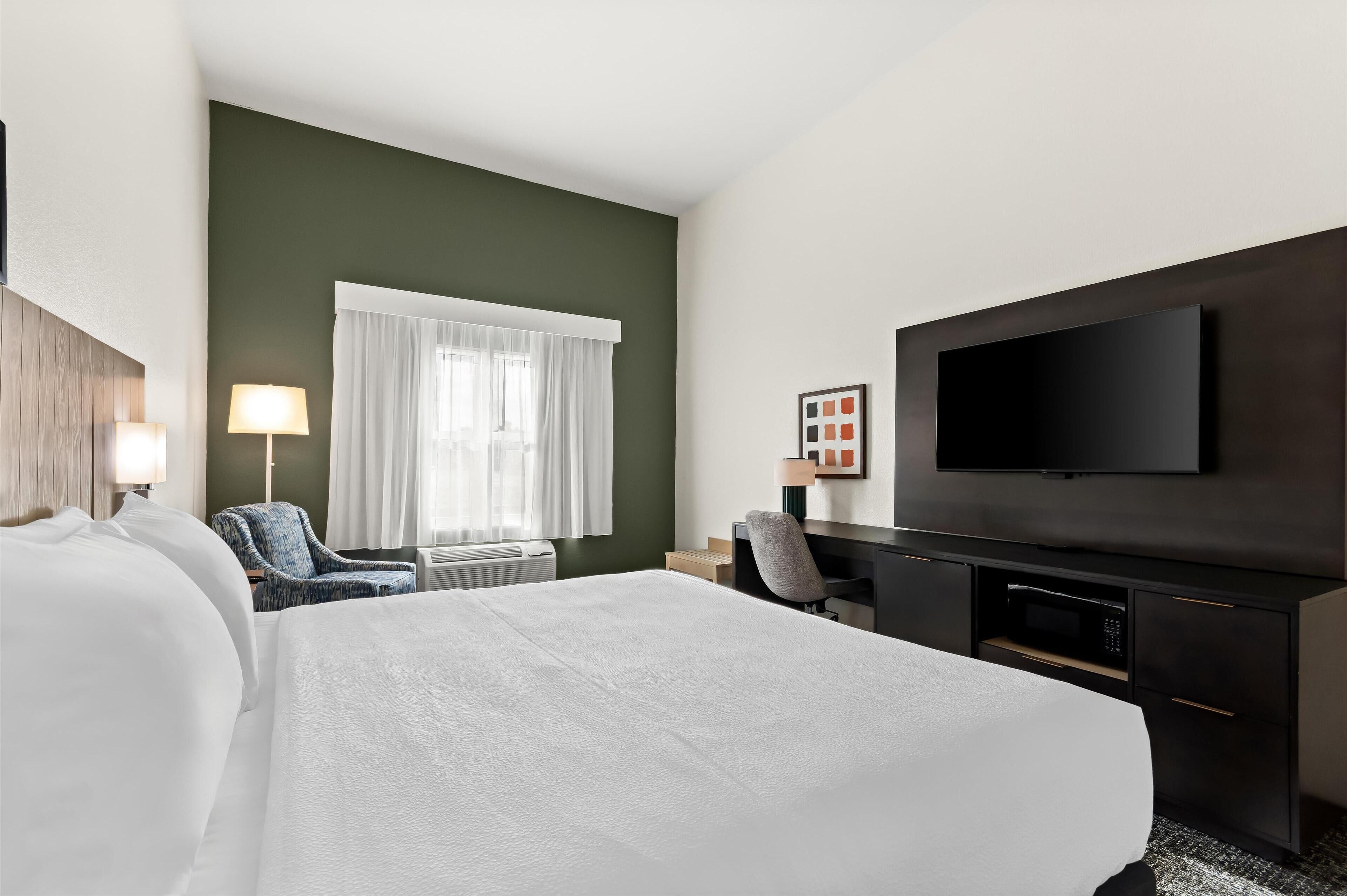 A hotel room with a large bed, a desk with a flat-screen TV, a chair, and a window with curtains.