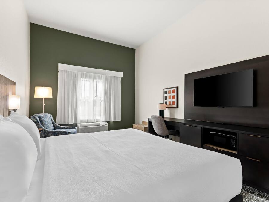 A hotel room with a large bed, a desk with a flat-screen TV, a chair, and a window with curtains.