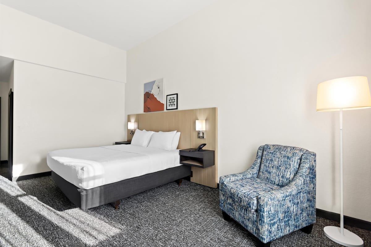 Quarto de hotel com cama, uma cadeira, luminária e mesa de cabeceira com telefone.