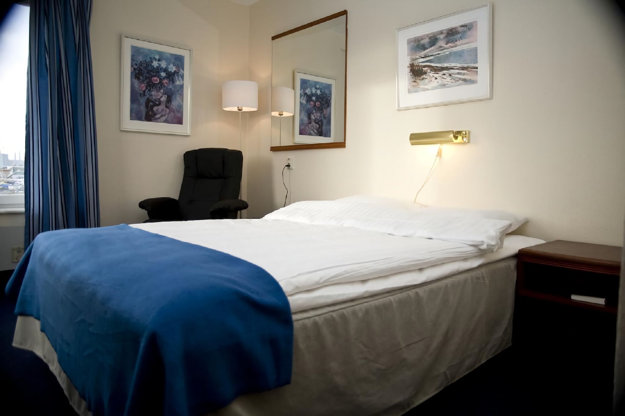 Una habitación de hotel con una cama, una silla, una mesita de noche y dos lámparas fijadas a la pared.
