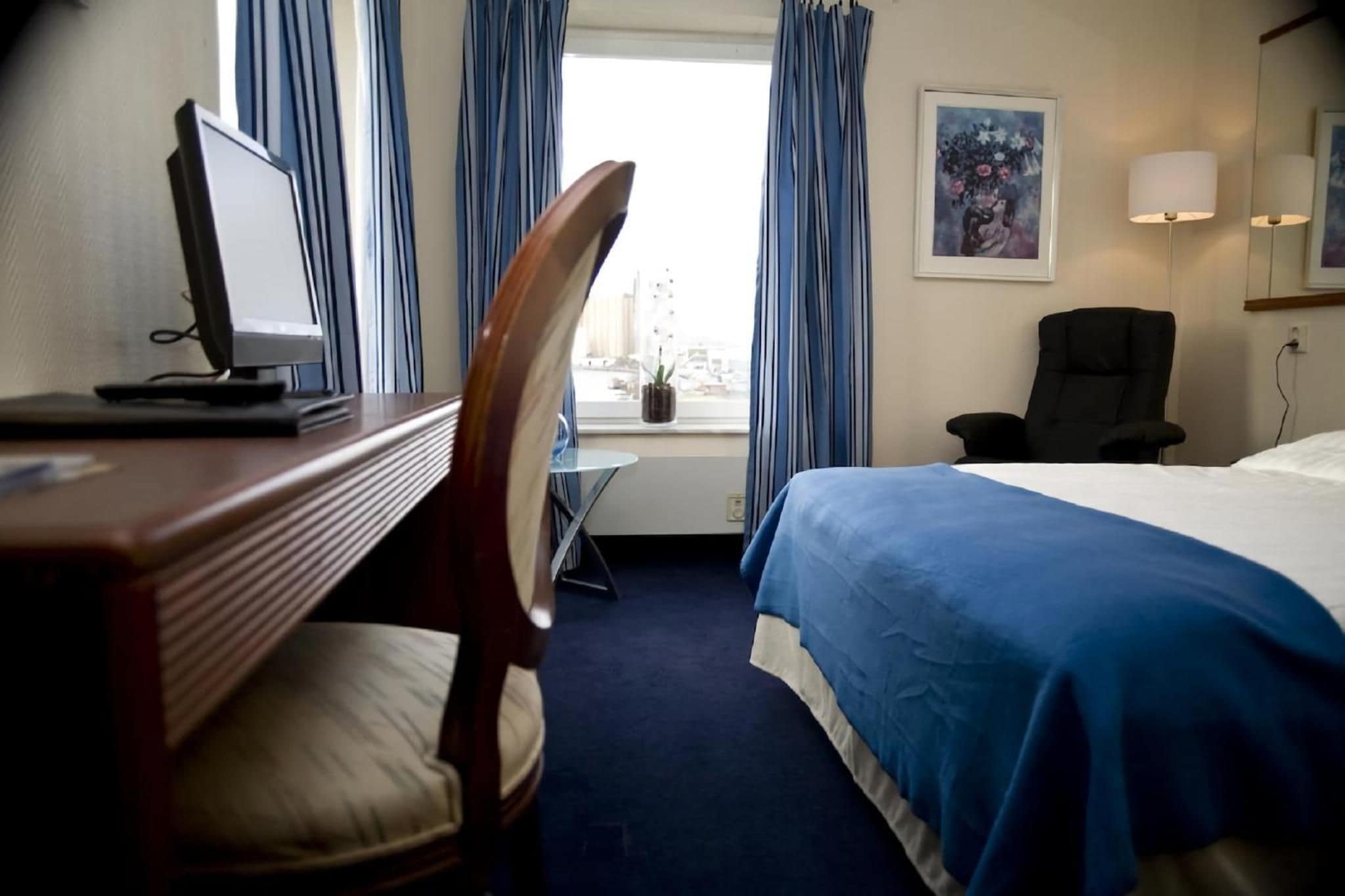 A hotel room with a bed, a desk with a computer, a chair, a window with curtains, and a painting on the wall.