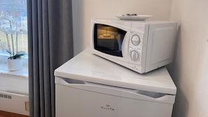 Un forno a microonde bianco su un elettrodomestico bianco, posizionato su un davanzale con vista su una pianta e una finestra con tende grigie.