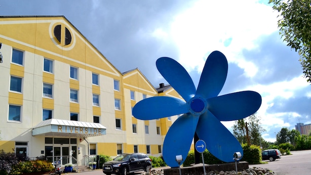 Un hotel con una grande scultura a forma di ventaglio blu di fronte, una facciata gialla e un cartello con la scritta 'RIVERSIDE'.