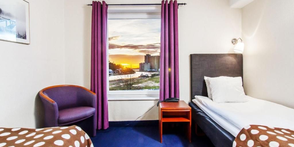 Ett hotellrum med en säng, en stol, ett litet bord och ett fönster med gardiner.