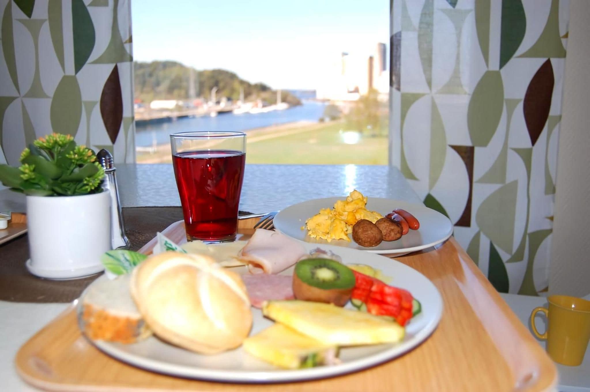 A breakfast tray with eggs, sausages, and fruit, a glass of red beverage, and a potted plant on a table by a window with a view of a body of water.