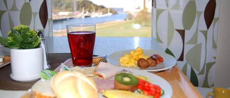 A breakfast tray with eggs, sausages, and fruit, a glass of red beverage, and a potted plant on a table by a window with a view of a body of water.