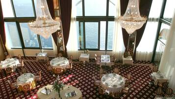 Un comedor con mesas redondas, sillas, candelabros y grandes ventanales con vista al agua.