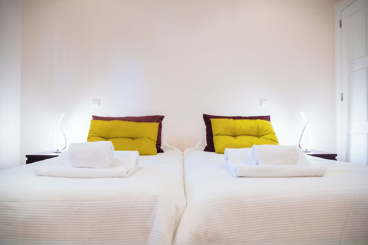 A hotel room with two beds, each with a white bedspread and two pillows, one yellow and one maroon. There are two bedside tables with lamps.