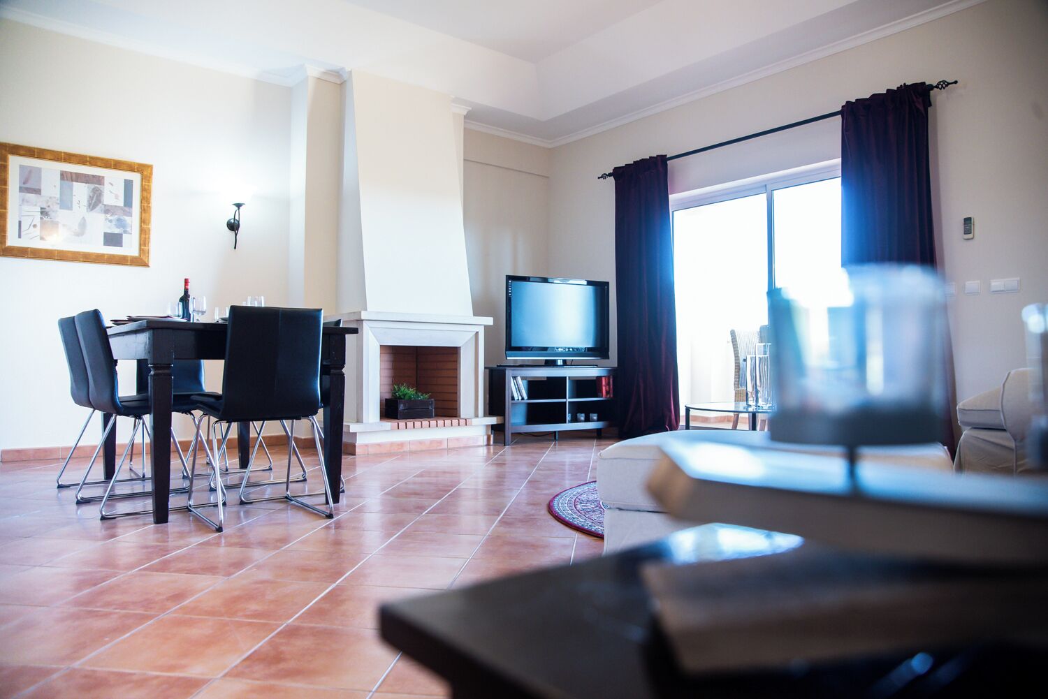 A living room with a fireplace, a dining area with chairs, a television, and a large window with curtains.