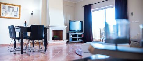 A living room with a fireplace, a dining area with chairs, a television, and a large window with curtains.