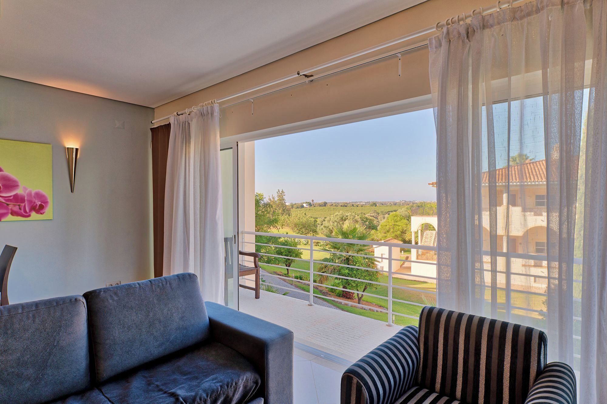 A balcony with a sofa and striped chair, a painting, and a view of greenery and buildings.