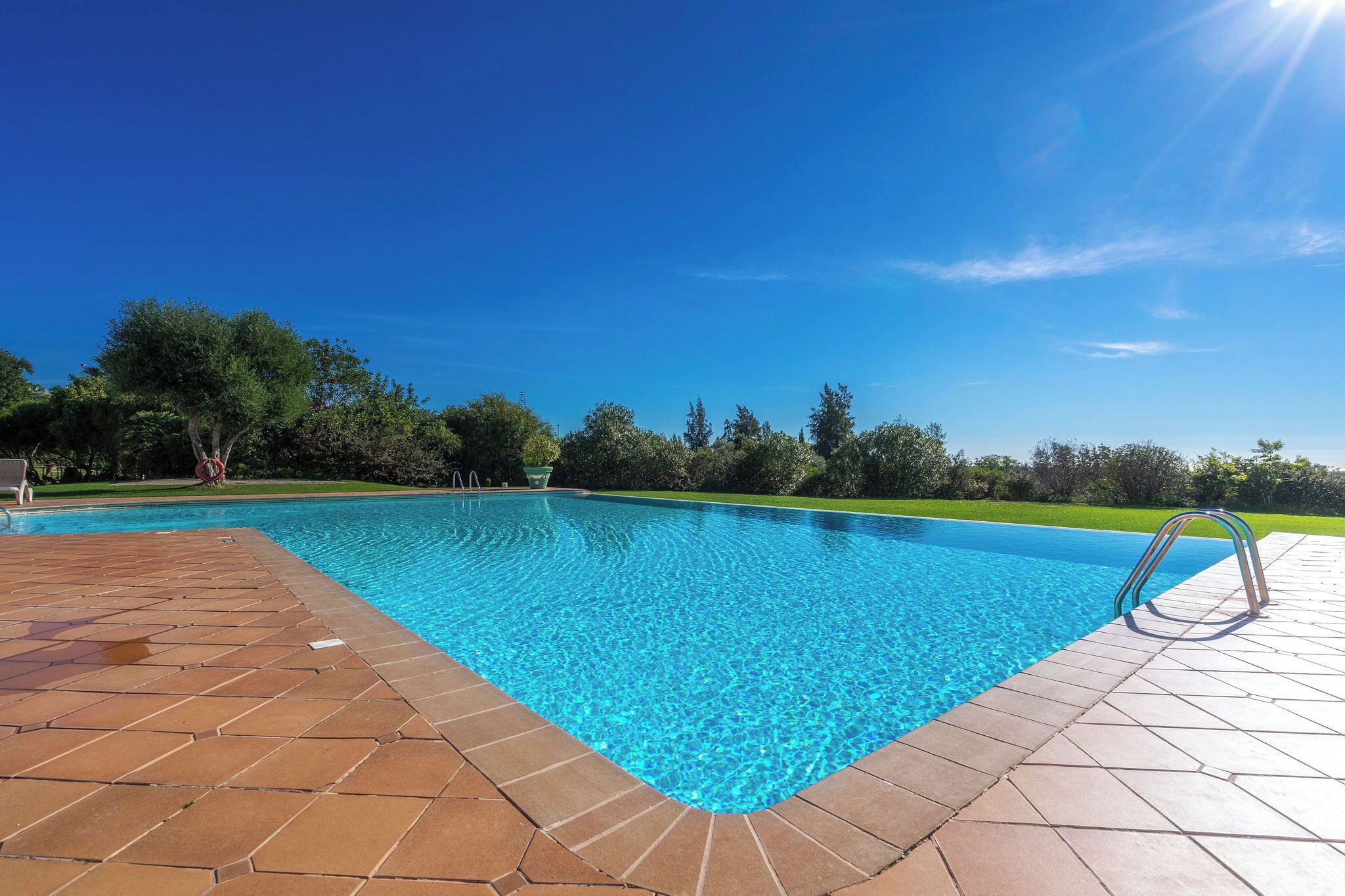 A clear blue swimming pool with a tiled deck, surrounded by trees and a clear sky.