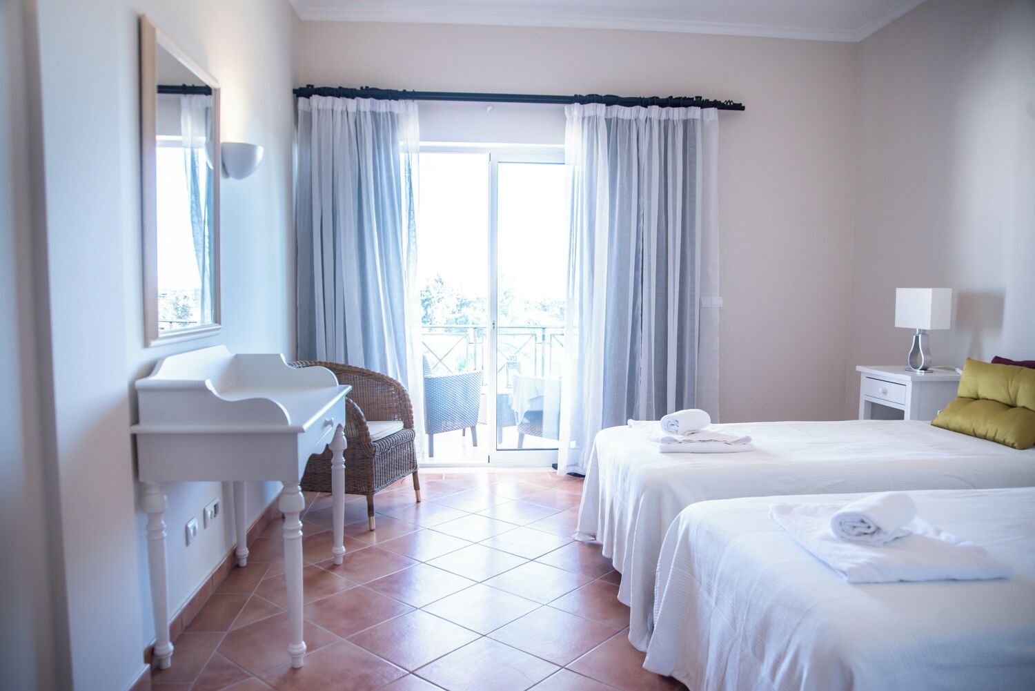 A hotel room with a bed, a desk, a chair, and a balcony with curtains.